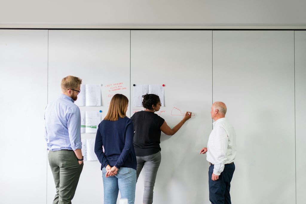 Vier Personen stehen an einer weißen Wand mit Notizen; eine Frau zeichnet mit rotem Stift ein Diagramm, die anderen schauen zu.