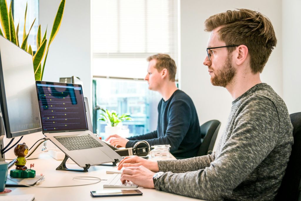 Ein Softwareentwickler mit Brille arbeitet konzentriert an einem Schreibtisch mit Laptop auf einem Ständer und externem Monitor; dahinter sitzt ein Kollege am Fenster.
