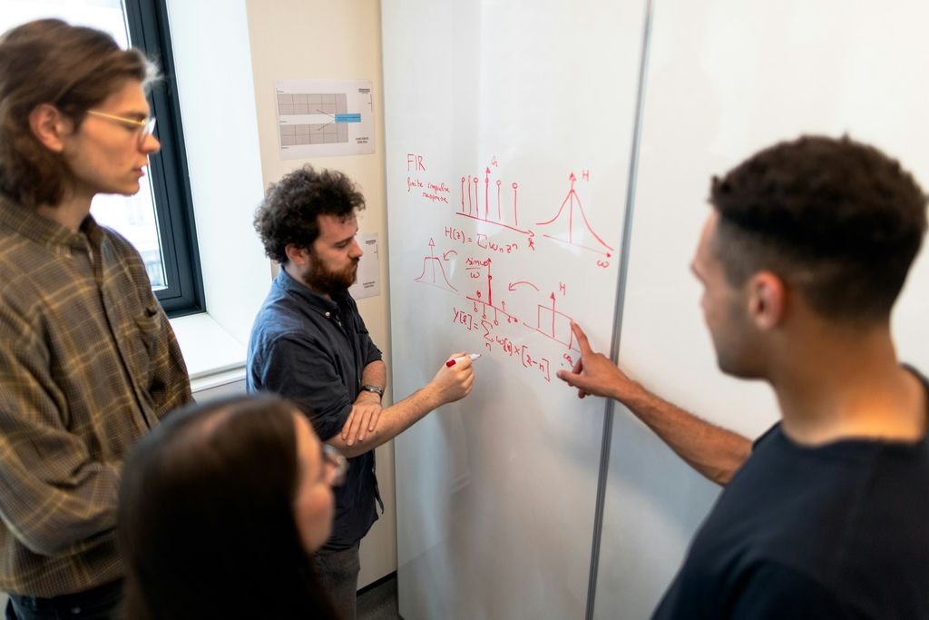 Vier Personen arbeiten an einem Whiteboard in einem hellen Büro. Ein Mann mit Bart schreibt mit rotem Stift Formeln und Signalverläufe an, ein Kollege zeigt auf eine Stelle, eine Frau und ein weiterer Mann sehen zu.