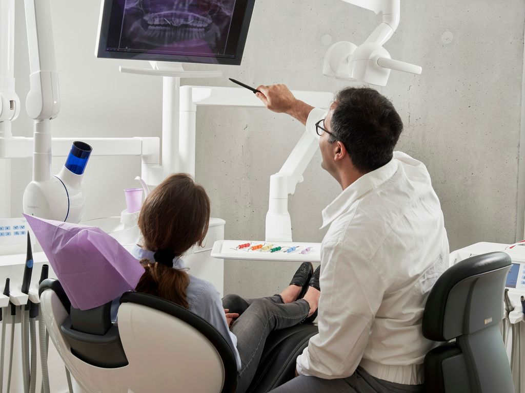 Ein Zahnarzt zeigt einer Patientin auf dem Behandlungsstuhl eine Röntgenaufnahme des Gebisses auf einem Monitor.