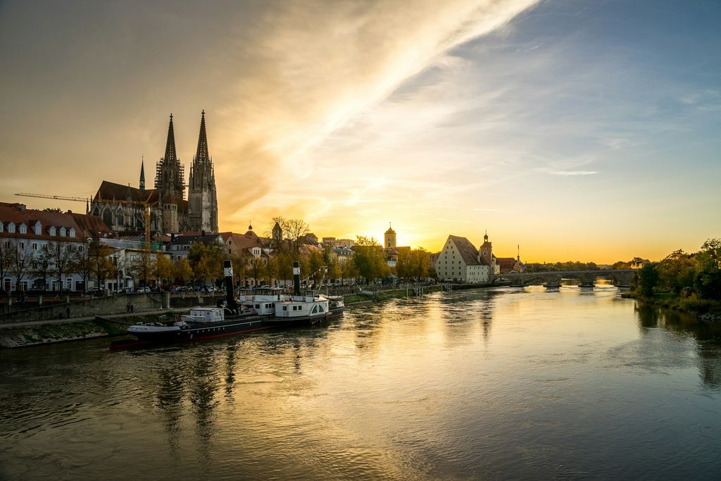 Regensburg bei Sonnenuntergang: der Dom St. Peter, die Altstadt und rechts die Steinerne Brücke über der Donau; am Ufer liegen Schiffe.