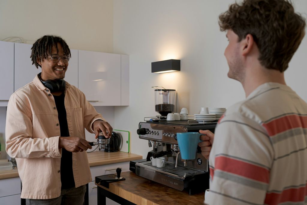 Zwei Kollegen machen Kaffeepause an einer Siebträgermaschine in einer Büroküche; einer lacht, der andere hält eine blaue Tasse.