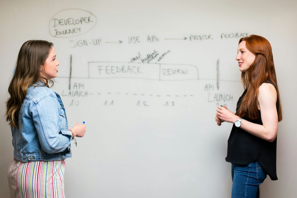 Zwei Frauen besprechen sich vor einem Whiteboard mit einer handgezeichneten Zeitleiste: 'Developer Journey', 'Sign-up', 'Use APIs', 'Feedback', 'Review' und 'API Launch'.